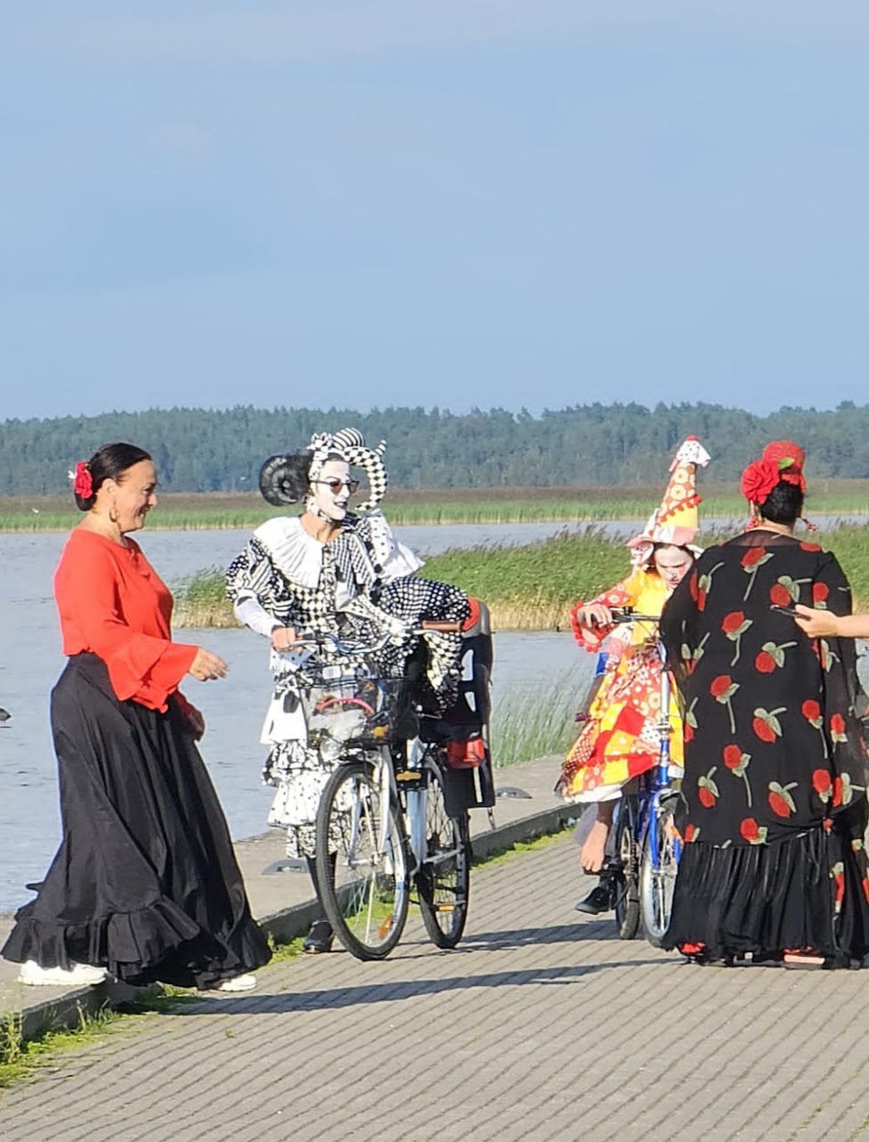 Pitkiin hameisiin pukeutuneita naisia meren rannalla. Yhden kasvot on maalattu valkoiseksi