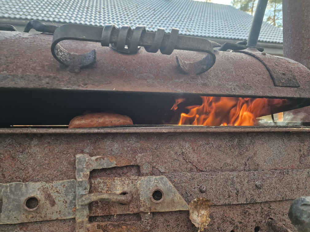 Grilliä koristaa raudasta taottu vyön solki. Grillin kansi on raollaan, ja raosta näkyy liekki ja makkaroita