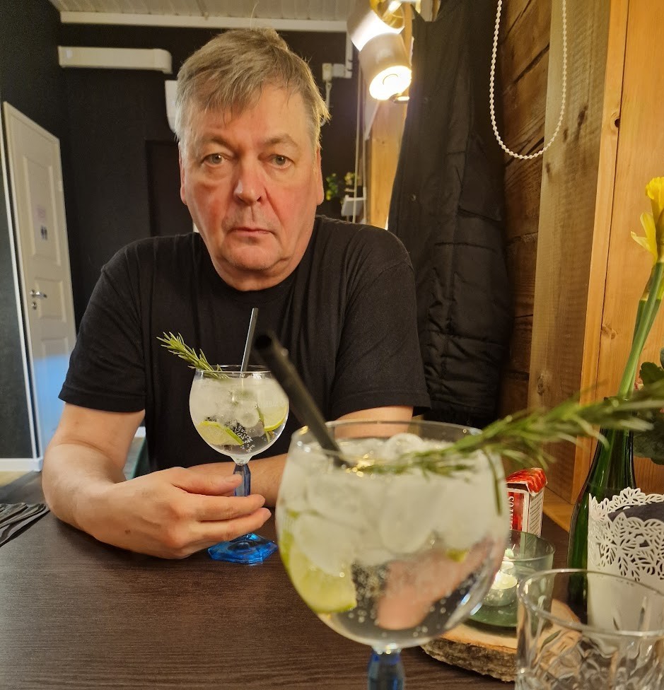 Ravintolan pöydässä kaksi rosmariinilla koristettua gin tonicia jalallisessa lasissa. Taustalla väsyneen näköinen mies