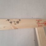 laudan päällä poikittaislauta ja naula, jonka päälle on piirretty kuva ja nimi